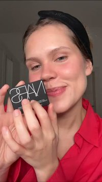 Compact NARS in una custodia nera opaca con logo bianco. Una persona lo tiene in mano, indossando una maglietta rossa e una fascia scura per capelli, in un ambiente neutro.