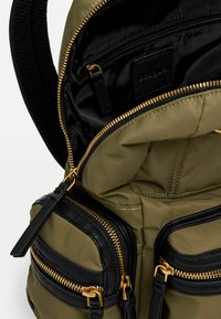 Zaino verde oliva con inserti in pelle nera, due cerniere frontali, accessori dorati, texture trapuntata e un ampio interno nero.
