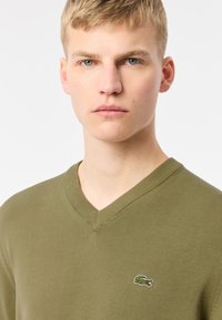 Suéter verde oliva de cuello en V hecho de tejido de punto. Presenta un cuello acanalado y un pequeño logo bordado en el pecho.
