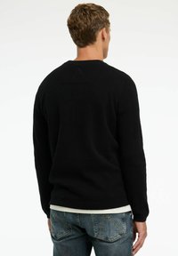 Schwarzer gerippter Strickpullover mit Rundhalsausschnitt, langen Ärmeln und strukturierten Details, getragen über einem weißen Hemd und kombiniert mit ausgewaschenen blauen Jeans.