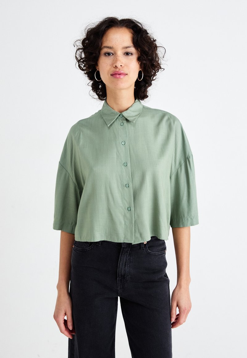 Camisa curta verde clara com frente abotoada, mangas curtas e colarinho clássico. Textura suave, com botões combinando.