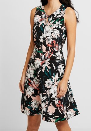 Frau trägt ärmelloses schwarzes Kleid mit weißem, pinkem und grünem Blumenmuster und kleinem Schlüssellochausschnitt am Dekolleté.