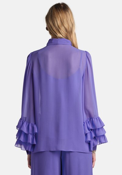 Lilla chiffonbluse med lange, puffede ærmer, der har lagdelte flæser ved manchetterne. Det gennemsigtige stof giver et let og flydende udseende.