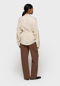 Lichtbeige overhemd met een gathered taille, lange mouwen en een ronde zoom, gecombineerd met losse, donkerbruine broeken en zwarte schoenen.
