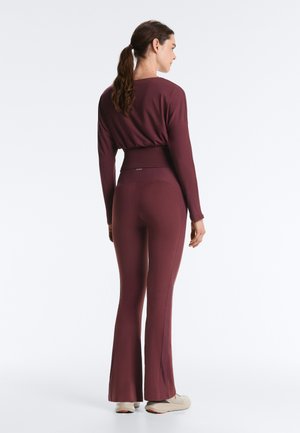 Donna in piedi di spalle che indossa un completo bordeaux composto da una maglia a maniche lunghe abbinata a pantaloni svasati e scarpe beige, su uno sfondo semplice.