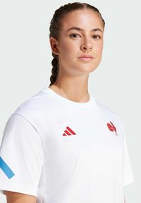 Camiseta blanca con el logo rojo de Adidas y emblema de gallo. Diseño de cuello redondo con textura suave. Raya de acento azul en la manga.