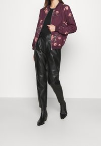 Burgundské bomber jacket s růžovou květinovou výšivkou, spárované s černými koženými kalhotami s vysokým pasem a černými lodičkami na vysokém podpatku.