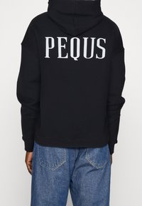 Felpa nera con cappuccio che presenta la scritta "PEQUS" in lettere bianche in grassetto sul retro. Vestibilità complessiva rilassata con polsini e orlo a coste.