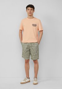 Lichtoranje katoenen T-shirt met zwarte grafische tekst, gecombineerd met groene geblokte shorts en beige sneakers met bruine accenten.
