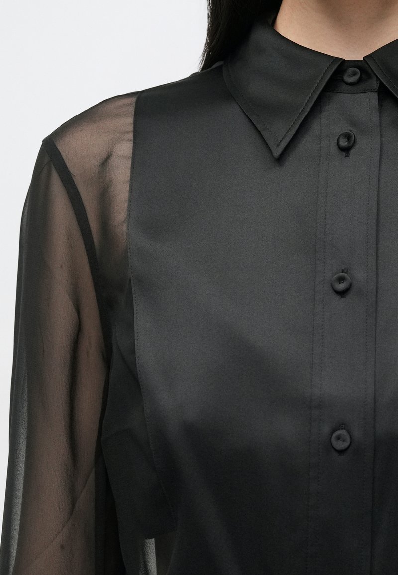 Chemise noire à boutons avec un front en tissu lisse et des manches transparentes et texturées ; elle comprend un col classique et une patte de boutonnage visible.