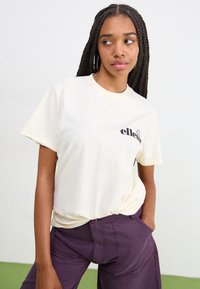 Krémové oversized tričko z bavlny s čiernym logom na ľavej strane. Krátke rukávy a okrúhly výstrih. Model skombinoval s fialovými nohavicami.