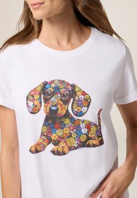 Vrouw in een wit T-shirt met een kleurrijke bloemenpatroon van een teckel op de voorkant, staand tegen een lichte achtergrond.