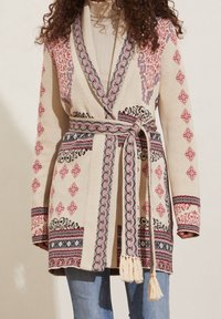 Stickad cardigan i beige med intrikata röda, blå och svarta mönster. Inkluderar ett matchande bälte med fransar och design med sjalkrage.