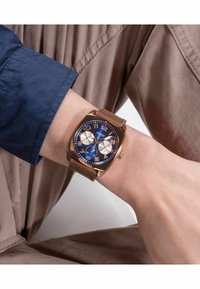 Reloj cuadrado con una esfera azul, acentos en tono dorado y tres subesferas; cuenta con una correa de ante marrón. Lleva puesto en una muñeca sobre un fondo neutro.
