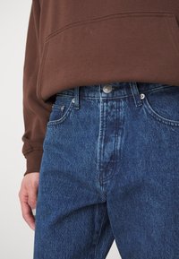 Denimjeans i en middels blå vask med høy midje, med knappestolpe og klassisk fem-lomme-design, kombinert med en brun hettegenser.