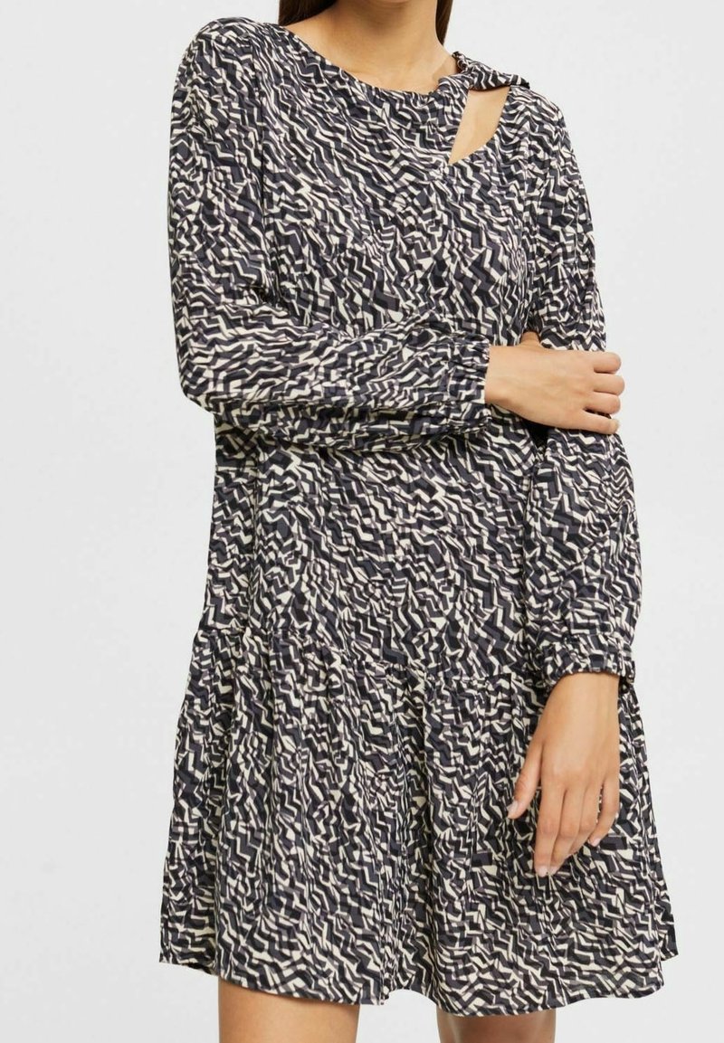 Femme portant une robe à manches longues à motif zigzag noir et blanc, avec une petite ouverture à l'épaule et une jupe à volants.