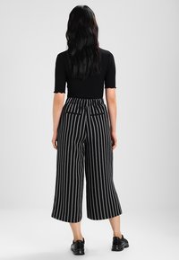 Zwarte cropped broek met verticale witte strepen, met een hoge tailleband, twee achterzakken en een losse pasvorm. Ggedragen met een strak zwart top.
