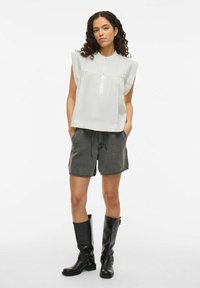 Witte mouwloze top met plooidetail, twee knopen bij de halslijn, gecombineerd met donkergrijze shorts en zwarte leren laarzen tot de knie.