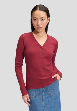 Ung kvinde med glat sort hår iført en langærmet rød ribbet top og blå denim nederdel mod en ensfarvet grå baggrund.