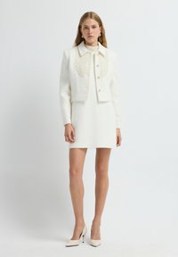 Chaqueta blanca texturizada con un gran detalle estampado y tres botones, combinada con un vestido blanco ajustado y zapatos de tacón beige.