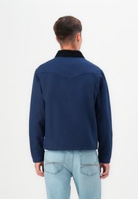 Chaqueta azul marino con cuello de pana, diseño simple, mangas largas y detalle de yugo en la parte posterior. Combinada con jeans de mezclilla azul claro.