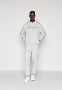 Γκρι σετ με φούτερ και παντελόνι φόρμας με μπροστινή τσέπη, που φέρει το λογότυπο "BJÖRN BORG" σε σκούρο πράσινο. Φοριέται με λευκά αθλητικά παπούτσια.