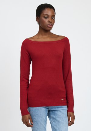Maglione rosso scivolato sulle spalle in morbido materiale a maglia con maniche lunghe e polsini a costina, abbinato a jeans azzurri. Logo dorato in accentuazione all'orlo.