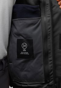 Černá bunda s kapsou na zip obsahující modrý pletený předmět. Má vnitřní štítek s nápisem "3-IN-1 SYSTEM" a pokyny pro údržbu.