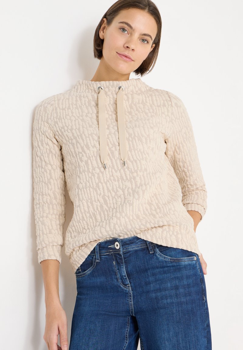 Kvinna med kort brunt hår som bär en texturerad beige långärmad tröja med dragsnören och blå jeans, stående mot en vit bakgrund.