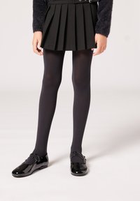 Gonna a pieghe nera, collant neri opachi e scarpe in vernice nera lucida con dettagli a cinturino. Design semplice e classico.