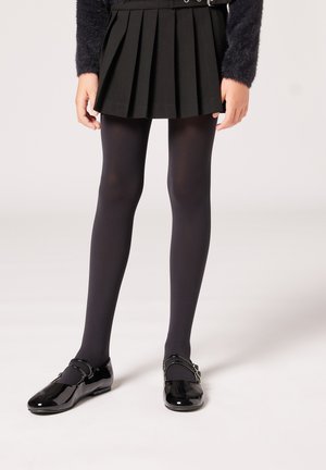 Schwarzer, gefalteter Rock, schwarze blickdichte Strumpfhosen und glänzende schwarze Lacklederschuhe mit einem Riemendetail. Einfaches und klassisches Design.