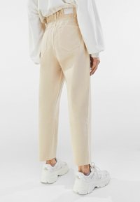 Person som bär beige höga croppade byxor med elastisk midja och vita klumpiga sneakers, stående mot en enkel vit bakgrund.