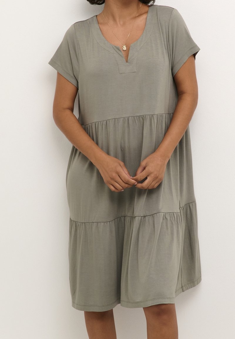 Femme portant une robe ample, à manches courtes, à volants, couleur sauge, avec un décolleté en encoches et un collier pendentif en or, debout devant un fond blanc.