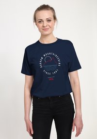 Młoda kobieta w granatowym t-shircie z grafiką gór i fal oraz napisem "Durham Mountaineering Since 1981" na przodzie.