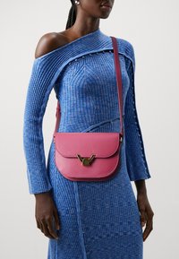 Borsa a tracolla in pelle rosa con chiusura a pattina, caratterizzata da dettagli in metallo dorato. Indossata sopra un abito blu a coste con bottoni decorativi.