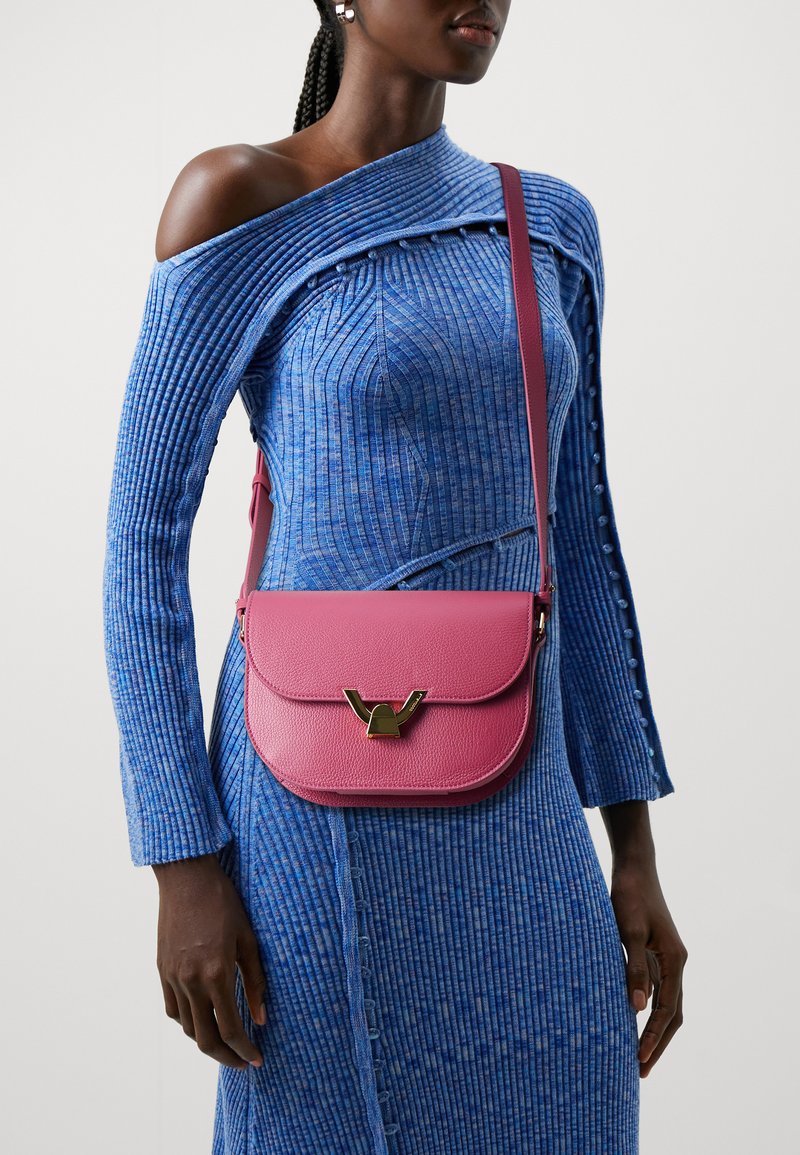 Borsa a tracolla in pelle rosa con chiusura a pattina, caratterizzata da dettagli in metallo dorato. Indossata sopra un abito blu a coste con bottoni decorativi.