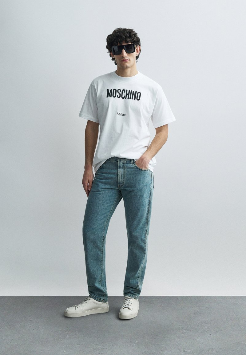Hvid t-shirt med "MOSCHINO" og "Milano" trykt i sort, kombineret med lyseblå jeans og hvide sneakers på en grå baggrund.