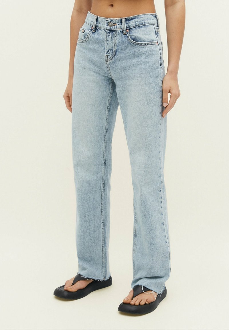 Jeans de mezclilla de tiro alto y pierna recta en azul claro, con un dobladillo sin rematar y un sutil desvanecimiento. Combinados con sandalias negras sin ajuste.