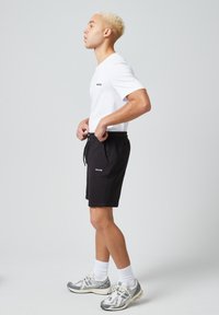 Witte T-shirt met korte mouwen en zwart logo, gecombineerd met zwarte shorts met trekkoord. Model draagt grijze sportschoenen met witte enkelkousen.