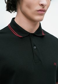 Close-up of person wearing black polo shirt with red double stripes on collar and small red logo on chest.