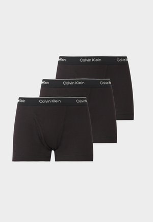 Muskuløs mand der står barfodet og bærer sorte Calvin Klein boxer briefs mod en ensfarvet lys grå baggrund.
