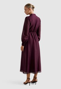 Dlhé vínovočervené šaty so zúženým pásom, nadýchanými rukávmi a golierom. Priehľadná textúra s plynulou sukňou. Modelka má na sebe čierne sandále na podpätku s remienkami.