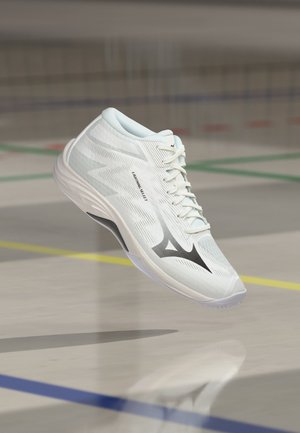 Chaussure de sport blanche avec logo noir et texte "Lightning Select", en l’air au-dessus du sol d’un terrain intérieur avec des lignes colorées.