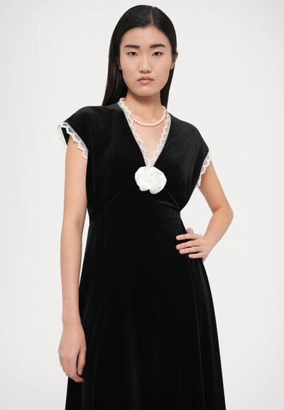 Vestido de veludo preto com decote em V, acabamento em renda nas mangas e um detalhe floral branco ao centro. A modelo posa com uma mão na anca.