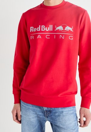 Mężczyzna w czerwonej bluzie z długim rękawem z napisem i logo „"Red Bull Racing"”, zestawionej z jasnoniebieskimi dżinsami.