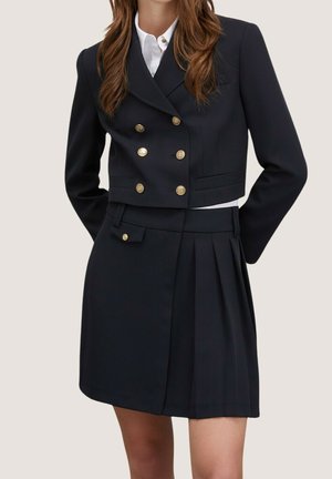 Vrouw draagt een op maat gemaakte zwarte cropped double-breasted blazer met gouden knopen en een bijpassende hoge taille geplooide rok.