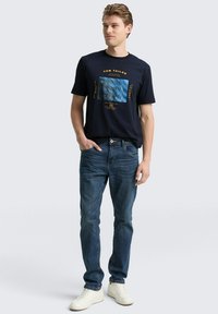 Granatowa koszulka z krótkim rękawem z niebieską grafiką i złotym napisem, w połączeniu z niebieskimi dżinsami i białymi sneakersami, prezentująca swobodny styl.