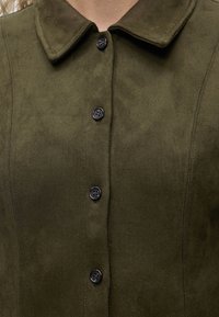 Gros plan d'une veste en daim verte avec un col et quatre boutons noirs visibles à l'avant.