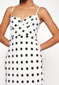 Vitt klänning med svarta prickar, tunna axelremmar, figurnära bodice, bärs av person med händerna på höfterna, på enfärgat vit bakgrund.