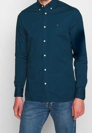 Homme portant une chemise à manches longues bleu sarcelle foncé boutonnée et un jean bleu, debout devant un fond clair uni.